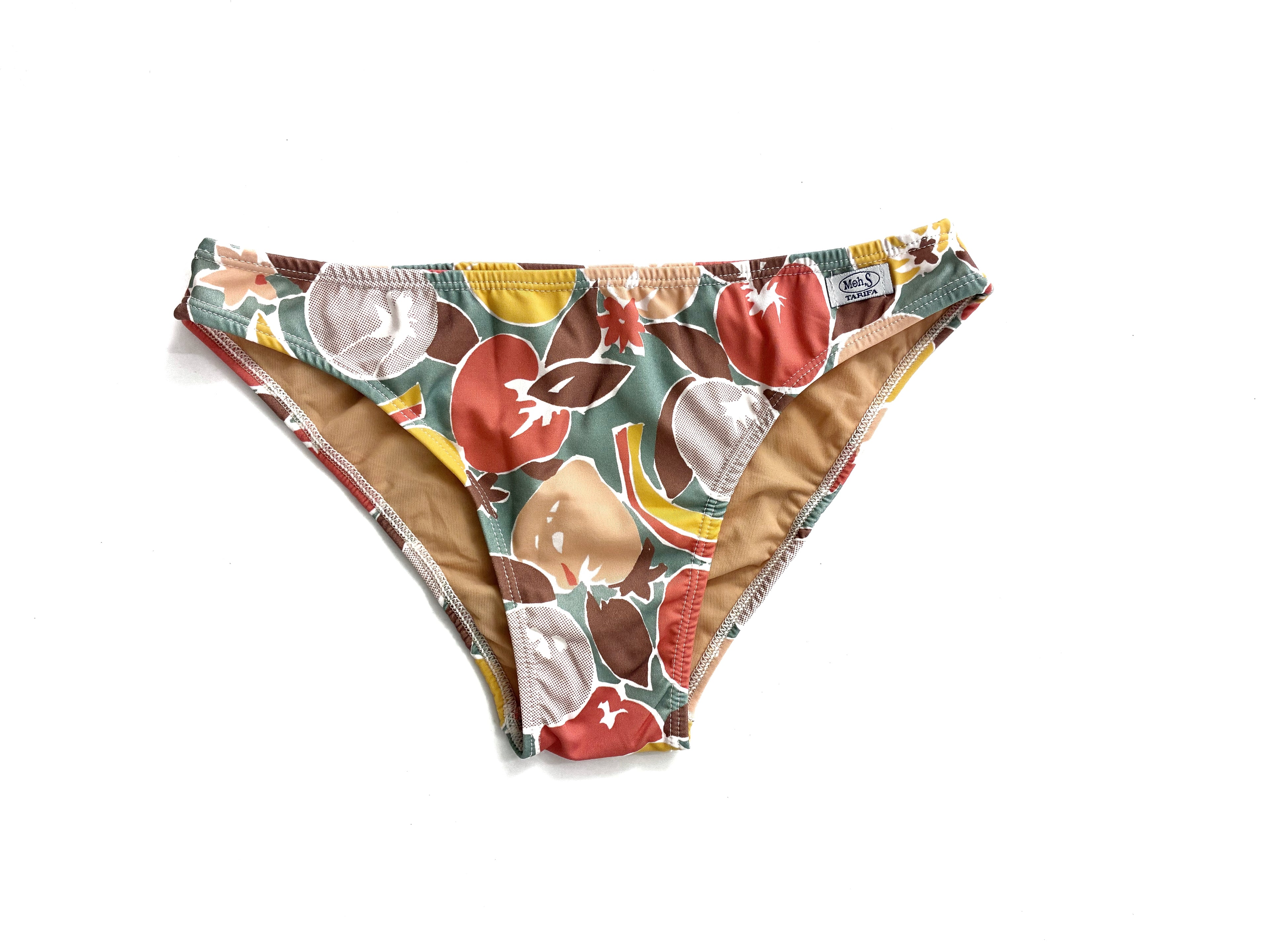 Braguita de bikini artesanal con estampado de frutas y verduras sobre fondo verde oliva en la playa.