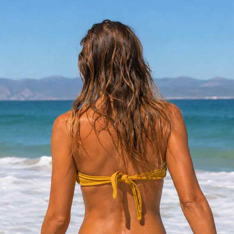 Modelo de espaldas con top de bikini bandeau artesanal en lurex dorado con tirantes finos al cuello.