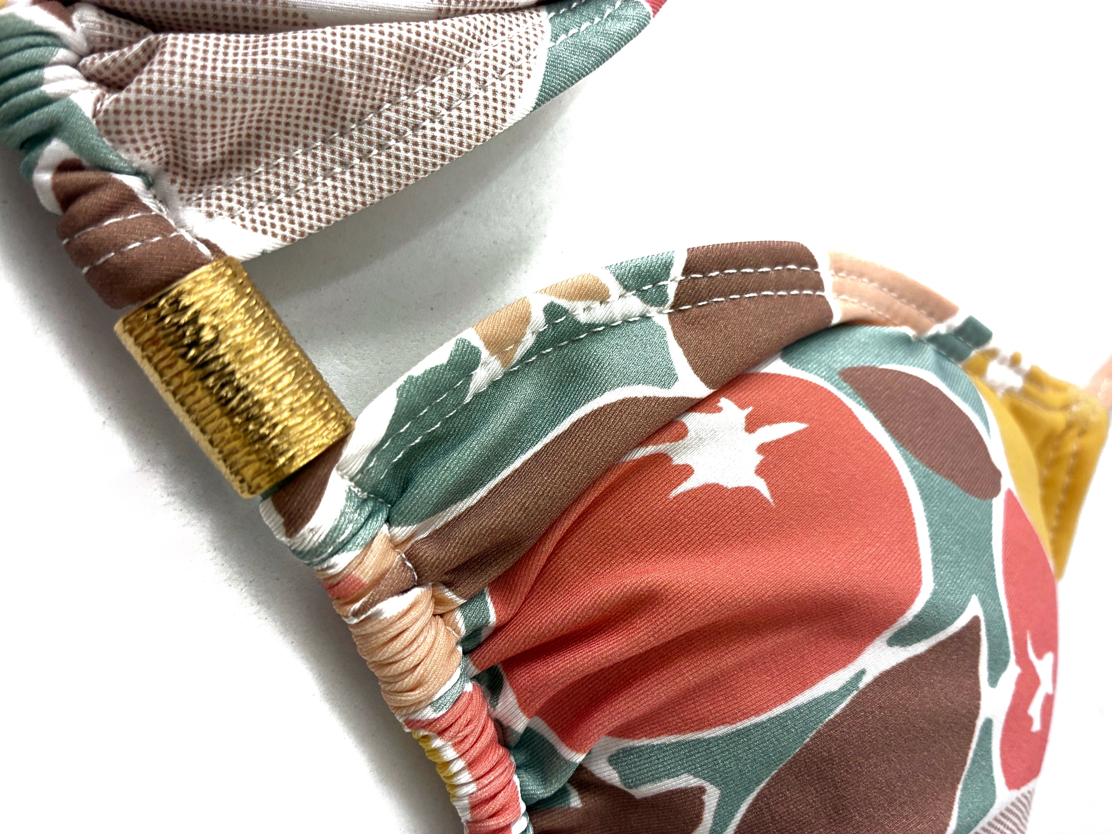Detalle dorado de top de bikini de triángulo cortina con estampado de frutas retro en tonos tierra, mostaza y verde con detalle de pieza dorada central.