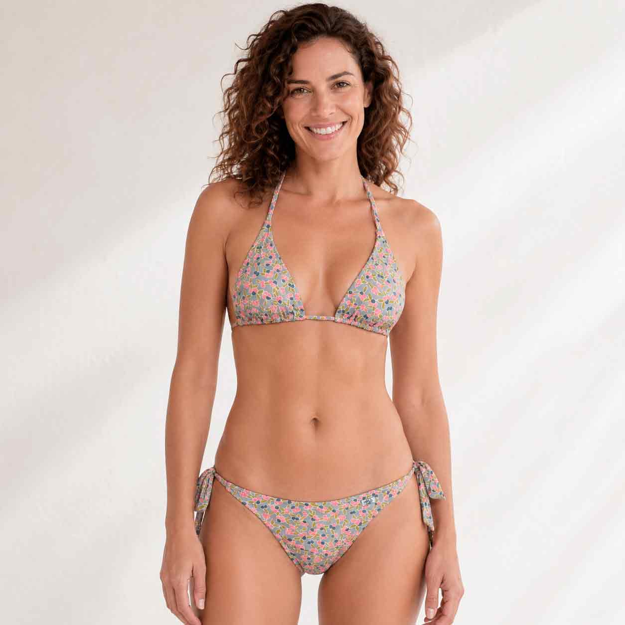 Mujer con top de bikini de triángulo con estampado de pequeñas flores y cerezas en tonos rosa, azul y verde sobre fondo gris claro.