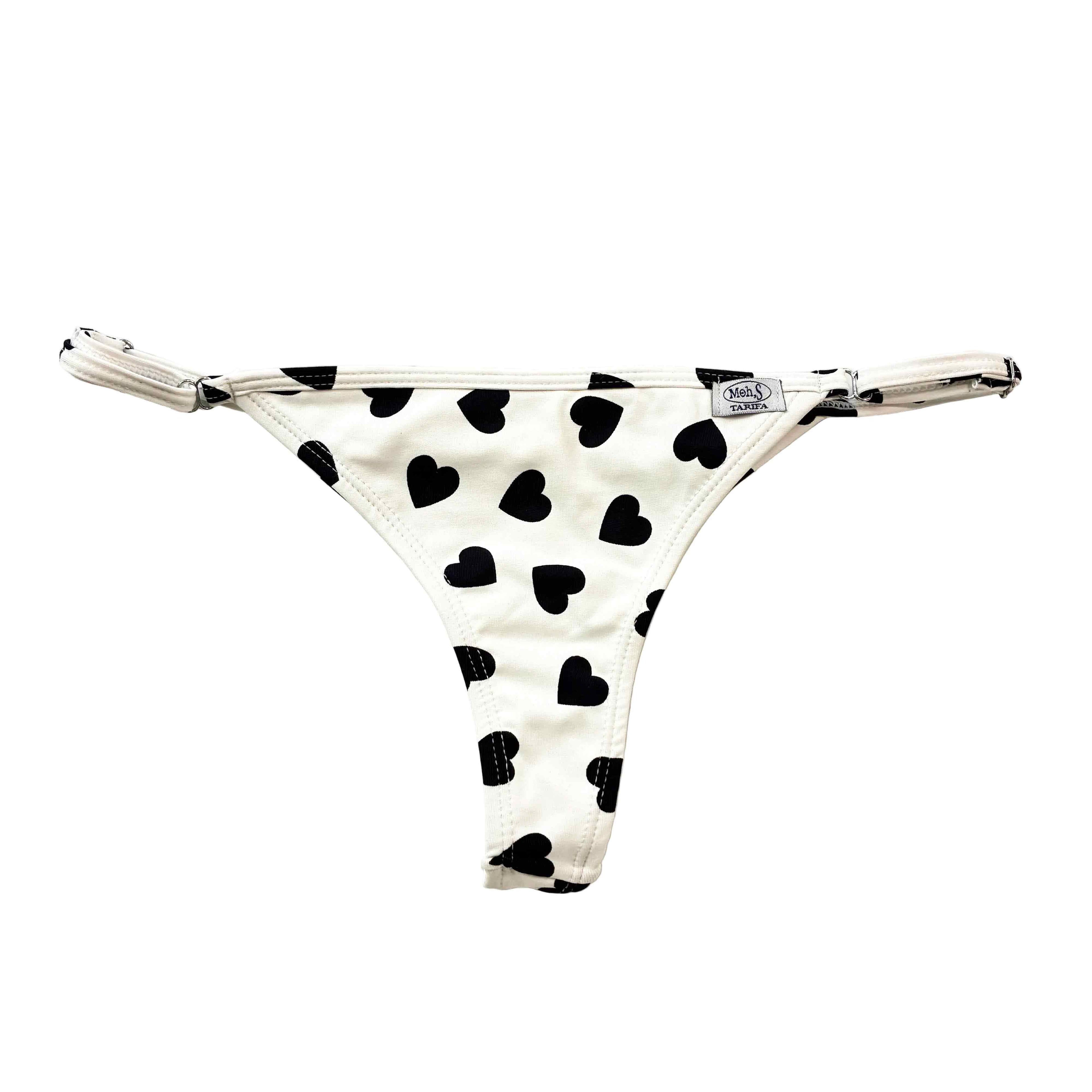 Tanga de bikini blanco con estampado de corazones negros y tirantes laterales regulables para un ajuste personalizado.