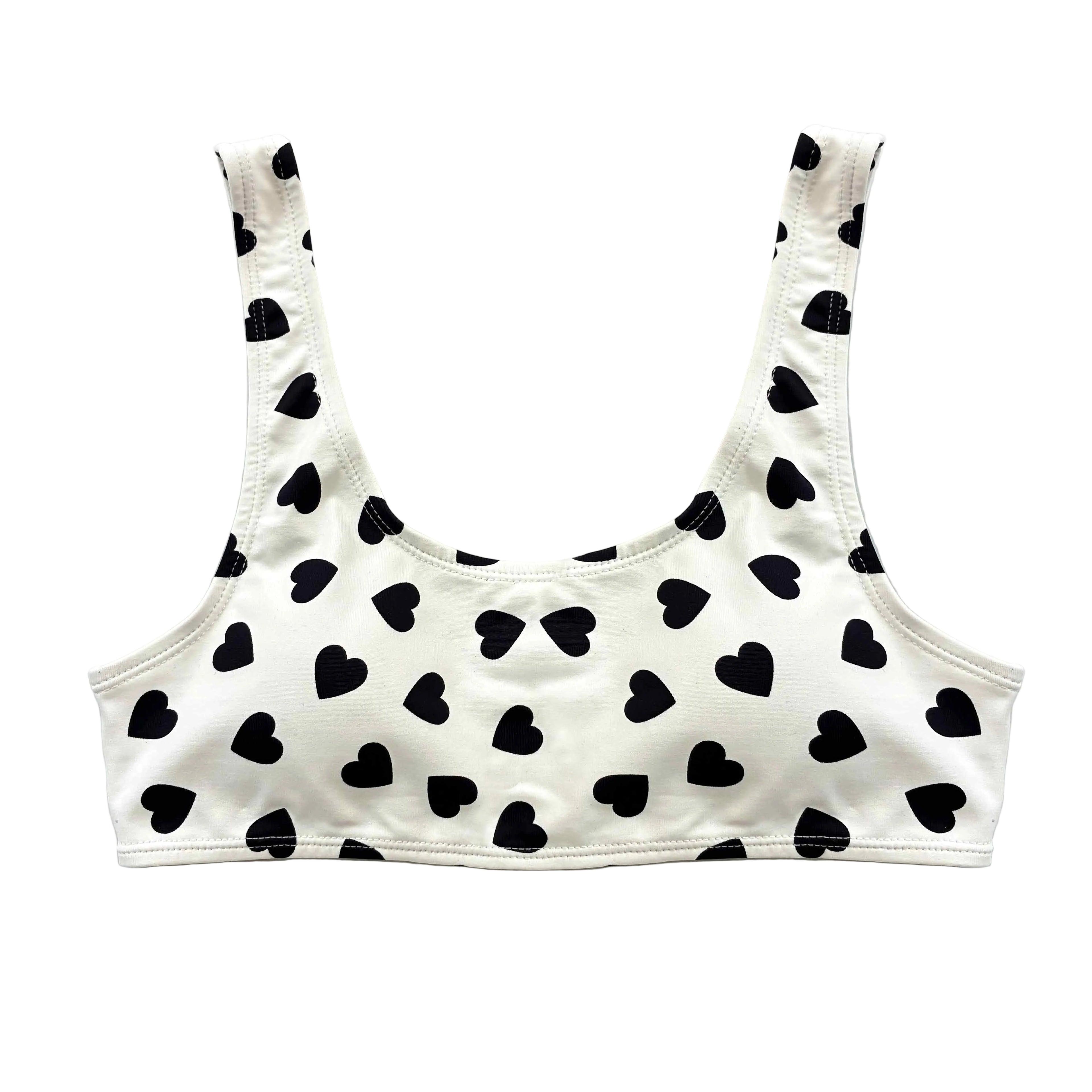 Top de bikini artesanal estilo deportivo en color blanco con estampado de corazones negros. Posee tirantes anchos y escote redondo, extendido sobre un fondo liso